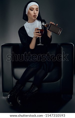 Full length shot of a nun, sitting on a chair. She's wearing dark nun's clothing and big boots. The nun is smoking, holding glass of wine in right hand and pour drink into glass. 