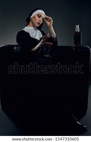 Full length shot of a nun, sitting on a chair. The nun is holding glass of wine in right hand, her left hand is on a forehead. There is a bottle with alcohol on a chair armrest.