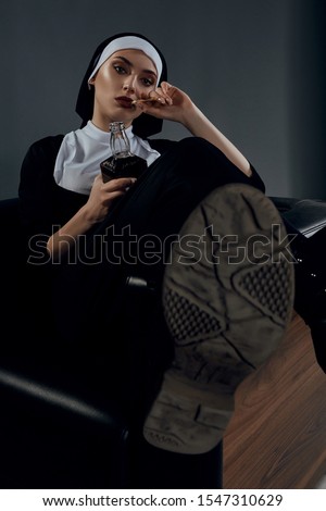 Full length shot of a nun, sitting on a black chair. There's a close-up of her soles in the frame. The nun is holding bottle of whiskey in right hand, her left hand is at her face. 