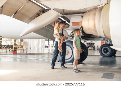 Full Length Shot Of Pilot In Uniform Showing Parts Of Airplane An Their Functions To Two Little Kids Who Came To Excursion At The Aircraft Hangar. Occupation, Childhood Concept