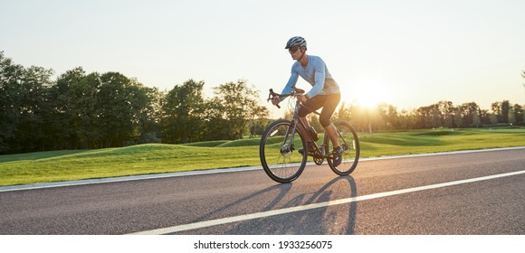 Full length shot of professional male racer in sportswear and helmet training, riding road bike on a cycle path in the park at sunset