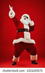 Full length shot of Santa Claus in sunglasses listening to music on headphones, singing and dancing. Merry Christmas. Party. Red background. 