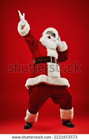 Full length shot of Santa Claus in sunglasses listening to music on headphones, singing and dancing. Merry Christmas. Party. Red background. 
