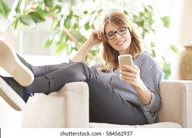 Full Length Shot Of A Smiling Mature Woman Reading Message On Her Mobile Phone While Sitting On Armchair At Home.