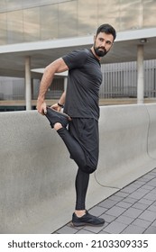 Full length shot of sporty adult bearded man stretches legs before running warms up prepares for workout dressed in sportswear looks away poses outdoors has muscular arms good physical condition