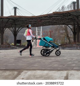 Full Length Shot Of Sporty Mom Running And Carrying A Stroller With A Child While Training In The Park On A Cold Autumn Day. Parenting, Healthy Lifestyle, Sports Concept. Side View