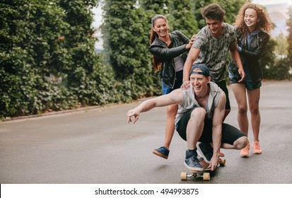 Full Length Shot Of Teenage Guys On Skateboard With Girls Pushing. Multi Ethnic Group Of Friends Having Fun Outdoors With Skateboard.