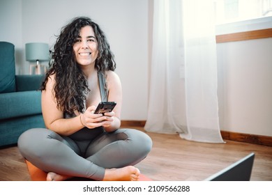 Full Length Shot Of Young Curvy Woman In Sportswear Sitting Floor Resting While Doing Fitness Yoga At Home. Using Phone Chatting. Losing Weight At Home. Online Class With The Laptop Concept.