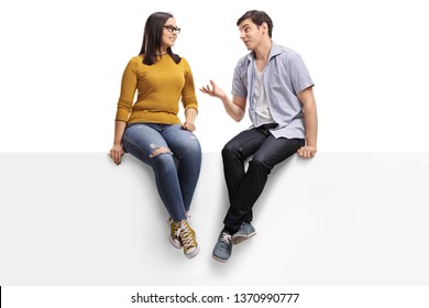 Full length shot of a young man sitting on a blank signboard and talking to a young woman isolated on white background
