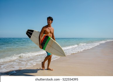 Full length shot of young man with surf board on beach. Handsome caucasian male holding surfboard walking on the sea shore and looking away. - Powered by Shutterstock