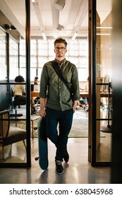 Full Length Shot Of Young Man With Skateboard Standing In Doorway Of Startup Office With People Working In Background.