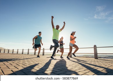 Full Length Shot Of Young People Running Along Seaside With Young Man Winning The Race.