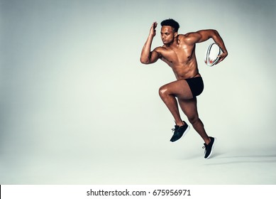 Full length shot of young rugby player training. Muscular male model running with rugby ball on grey background. - Powered by Shutterstock