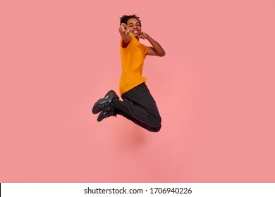 Full length shot of young surprised dark skinned man jumping in the air and pointing, choosing something, isolated on pink studio background. Freedom of choices, inspiration, human emotions concept.