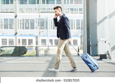 Full length side portrait of business man walking with luggage on telephone call at station