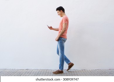 Full Length Side Portrait Of A Cheerful Young Man Walking On Sidewalk And Reading Text Message On Mobile Phone