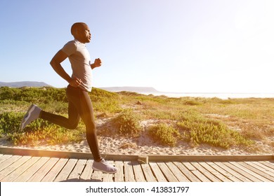 Full length side portrait of fitness man running on the boardwalk at the beach on a bright sunny day