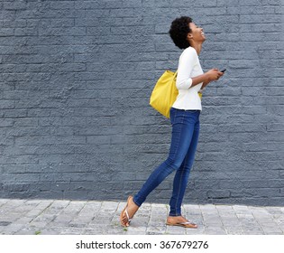 Full Length Side Portrait Of A Smiling Black Woman Walking On Street With Cell Phone