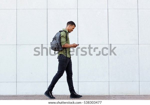 スマートフォンとヘッドフォンを使って歩く笑顔の男性の全長横のポートレート の写真素材 今すぐ編集