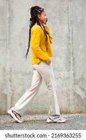 Full Length Side Portrait Of Stylish Young Black Woman Talking On Cellphone While Walking Against Gray Wall Outdoors