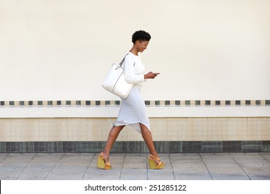 Full length side portrait of a young woman walking and sending text message on cell phone 