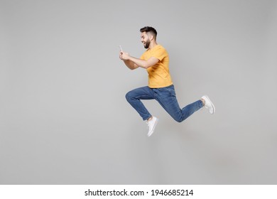 Full Length Side Profile View Of Young Bearded Cool Student Man 20s In Yellow T-shirt Jump High Hold Mobile Cell Phone Chatting Using Fast Internet Run Isolated On Grey Background Studio Portrait.