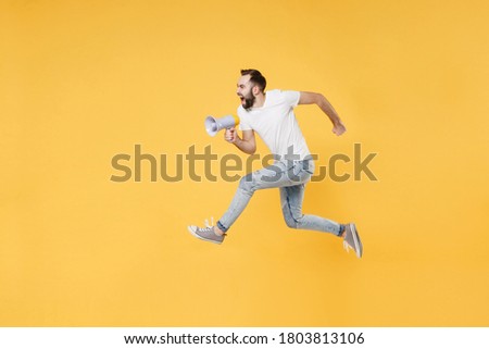 Full length side profile view image of crazy fun young bearded man 20s in white basic white t-shirt high jumping like running, screaming in megaphone isolated on yellow background, studio portrait.