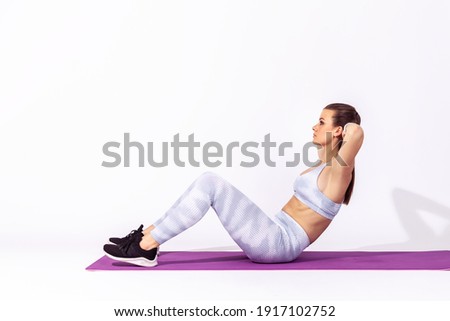 Full length side view athletic woman in white tights and top lying on mat and training doing sit ups, exercising abdominal muscles, lower-body workouts. Indoor studio shot isolated on gray background