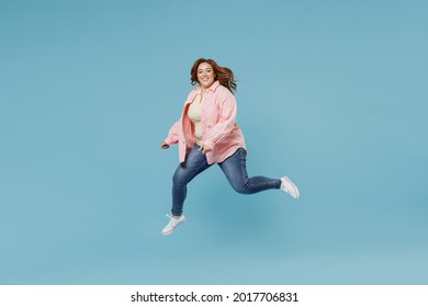 Full Length Side View Caucasian Young Redhead Chubby Overweight Woman 30s With Curly Hair In In Pink Shirt Jeans Casual Clothes Running Jump High Isolated On Pastel Blue Background Studio Portrait.