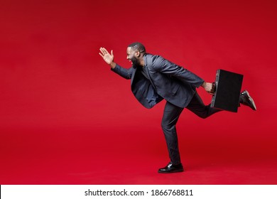 Full Length Side View Of Cheerful Young African American Business Man 20s In Classic Jacket Suit Hold In Hand Briefcase Running Looking Aside Isolated On Bright Red Color Background Studio Portrait