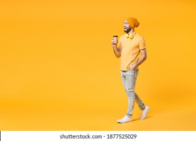 Full length side view of cheerful young bearded man 20s wearing basic casual t-shirt headphones hat hold paper cup of coffee or tea looking aside isolated on bright yellow background, studio portrait - Powered by Shutterstock