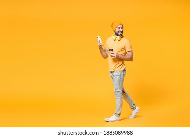 Full length side view of cheerful young man in basic casual t-shirt headphones hat using mobile cell phone hold paper cup of coffee or tea looking aside isolated on yellow background, studio portrait - Powered by Shutterstock