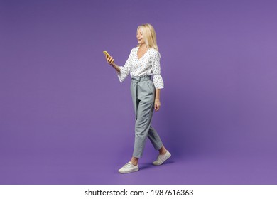 Full Length Side View Cheerful Laughing Elderly Gray-haired Blonde Woman Lady 40s 50s In White Dotted Blouse Using Mobile Cell Phone Typing Sms Message Isolated On Violet Background Studio Portrait