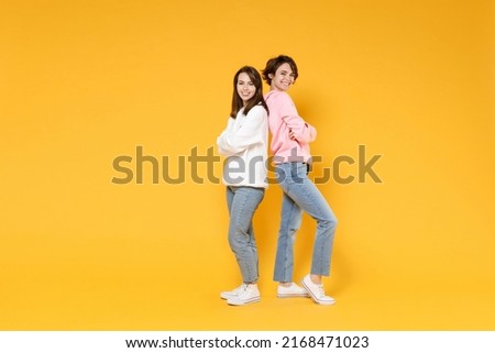 Full length side view of cheerful two young women friends 20s wearing casual white pink hoodies standing back to back holding hands crossed isolated on bright yellow color background studio portrait