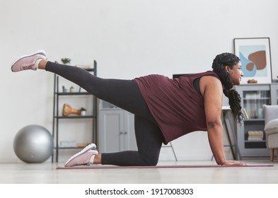 Full length side view at curvy African American woman working out at home and smiling while stretching on yoga mat, copy space