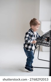 Full length side view of a cute toddler boy in a plaid shirt playing with a toy car on a black wire chair. Minimalist bright white studio background with plenty of copy space.