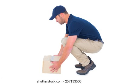Full length side view of delivery man crouching while picking cardboard box on white background