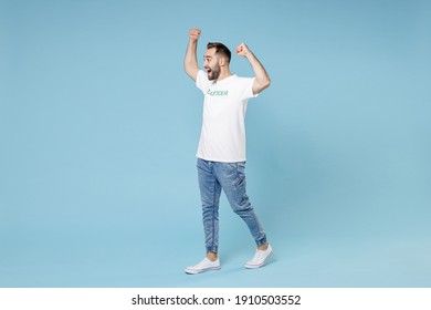 Full Length Side View Excited Young Man In White Volunteer T-shirt Clenching Fists Like Winner Isolated On Blue Background Studio Portrait. Voluntary Free Work Assistance Help Charity Grace Concept