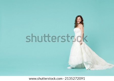 Full length side view of excited bride young woman 20s in beautiful white wedding dress casual gumshoes looking camera isolated on blue turquoise background studio. Ceremony celebration party concept
