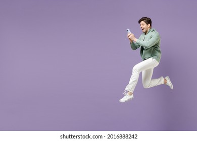 Full length side view fun smiling caucasian man in casual mint shirt white t-shirt using mobile cell phone chat online jump high isolated on purple background studio portrait People lifestyle concept