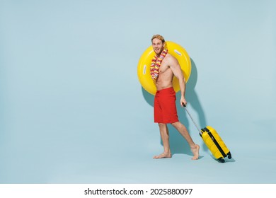Full length side view fun traveler tourist man in shorts swimsuit hawaii lei hold inflatable ring suitcase walk isolated on blue background. Passenger travel abroad weekend getaway. Air flight concept