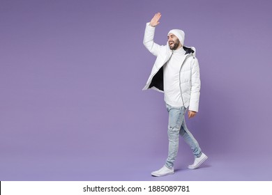 Full Length Side View Funny Man In White Windbreaker Jacket Hat Waving Greeting With Hand As Notices Someone Isolated On Purple Background Studio Portrait. People Lifestyle Cold Winter Season Concept