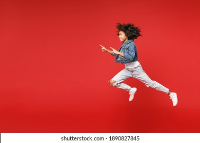 Full length side view of funny little african american kid girl 12-13 in denim jacket jumping pointing index fingers aside isolated on red background children portrait. Childhood lifestyle concept - Shutterstock ID 1890827845