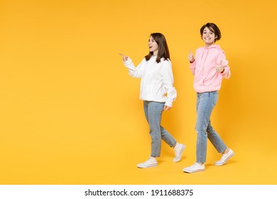 Vollständige Seitenansicht von lustigen zwei jungen Freundinnen 20ern, die beigefarbene, weiße rosafarbene Hoodies tragen, die Zeigefinger beiseite zeigen, die Daumen aufzeigt einzeln auf hellgelbem Hintergrund, Studioaufnahme