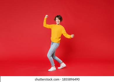 Full length side view of happy joyful laughing young brunette woman 20s wearing casual yellow sweater clenching fists doing winner gesture isolated on bright red colour background studio portrait