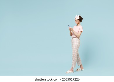 Full length side view happy young woman in pajamas jam sleep eye mask rest relaxing at home hold mobile cell phone walking going isolated on pastel blue background. Good mood night bedtime concept.