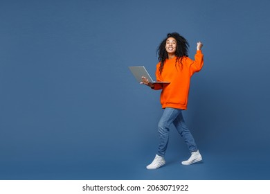 Full Length Side View Of Happy Young African American Woman Wearing Basic Bright Orange Sweatshirt Working On Laptop Pc Computer Doing Winner Gesture Isolated On Blue Color Background Studio Portrait