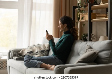 Full length side view happy millennial hispanic woman wearing wired earphones, choosing favorite audio tracks in cellphone playlist, playing mobile games or communicating in social networks at home. - Shutterstock ID 2107355399