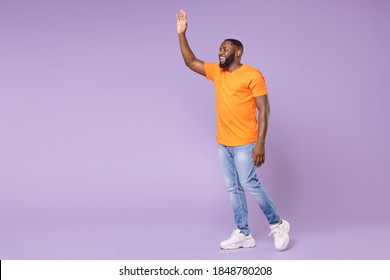 Full length side view of joyful young african american man wearing basic casual orange t-shirt waving and greeting with hand as notices someone isolated on pastel violet background, studio portrait