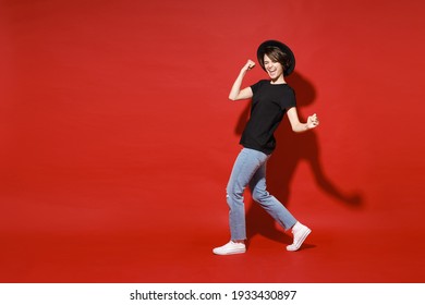 Full length side view of joyful young brunette woman 20s in casual black t-shirt hat doing winner gesture celebrating clenching fists say yes isolated on bright red color background studio portrait